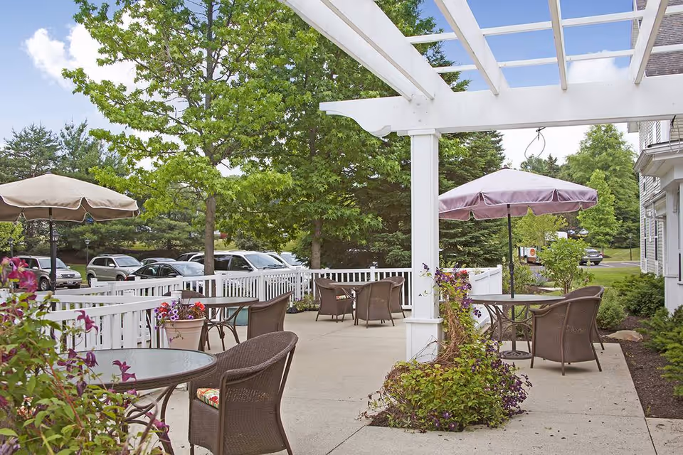 Outdoor patio with wicker tables and chairs under a white pergola and umbrellas, surrounded by potted plants and a white fence with parked cars beyond.