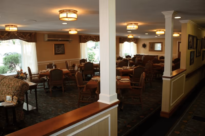 Interior view of a dining room in a senior living facility with multiple tables covered with brown tablecloths, set with plates and silverware. The room has several chairs around each table, large windows with curtains letting in natural light, decorative columns, and ceiling lights. There is a floral arrangement on a side table and framed pictures on the walls.