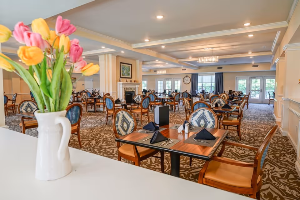 A spacious dining room with multiple wooden tables and chairs arranged neatly. Each table is set with black napkins, silverware, and salt and pepper shakers. The room features patterned carpeting, a fireplace, large windows with curtains, and ceiling lights. In the foreground, there is a white vase with colorful tulips.
