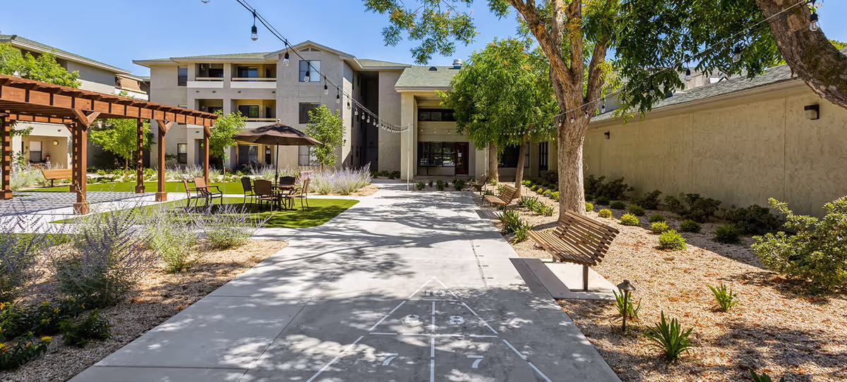 Outdoor courtyard area of a senior living facility with a concrete walkway featuring a shuffleboard game, wooden benches under trees, a pergola with seating, and a multi-story building in the background under a clear blue sky.