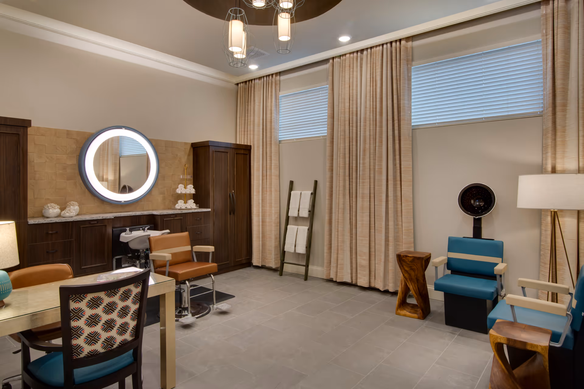A well-lit salon room with a hair washing station, a round illuminated mirror above a countertop with cabinets, two salon chairs, a ladder-style towel rack with white towels, and two blue salon chairs with a hair dryer hood. The room has beige curtains, tiled floor, and modern ceiling lights.