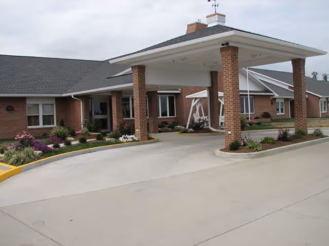 Exterior view of a single-story brick building with a covered entrance supported by brick columns. There are landscaped flower beds with various plants and flowers around the entrance area, and a white swing is visible under the covered area.