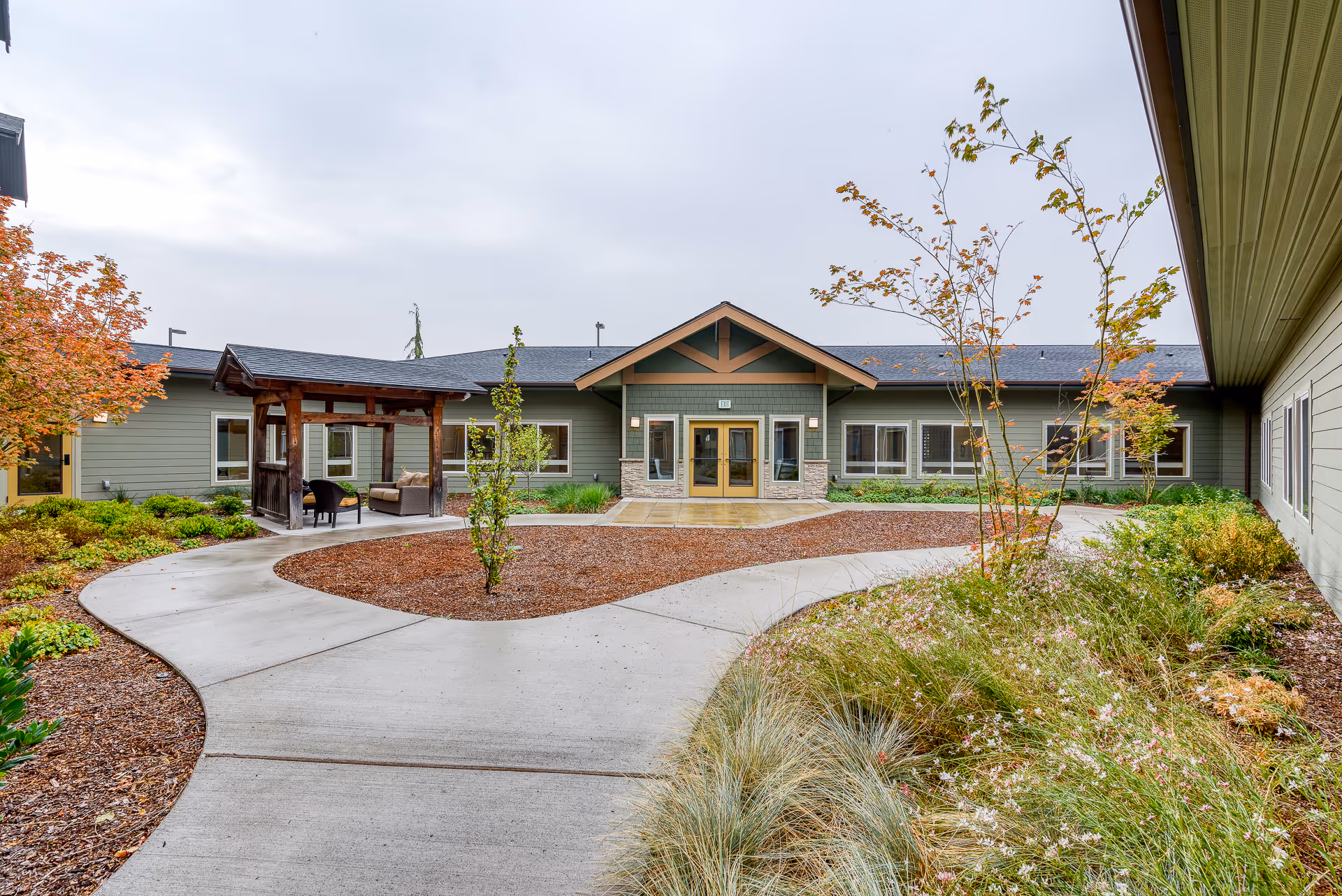 Outdoor courtyard area of Meridian at Stone Creek featuring a curved concrete walkway, landscaped garden beds with various plants and small trees, and a wooden pergola with seating. The building exterior is visible with green siding, multiple windows, and a central entrance with double doors under a peaked roof.