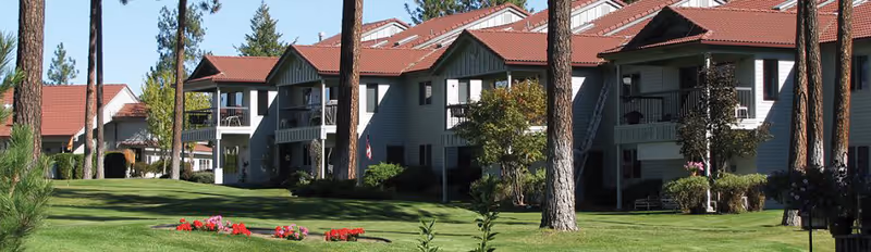 Exterior view of a retirement village building with multiple units, featuring gray siding and red roofs, surrounded by tall pine trees, green lawns, and flower beds under a clear blue sky.