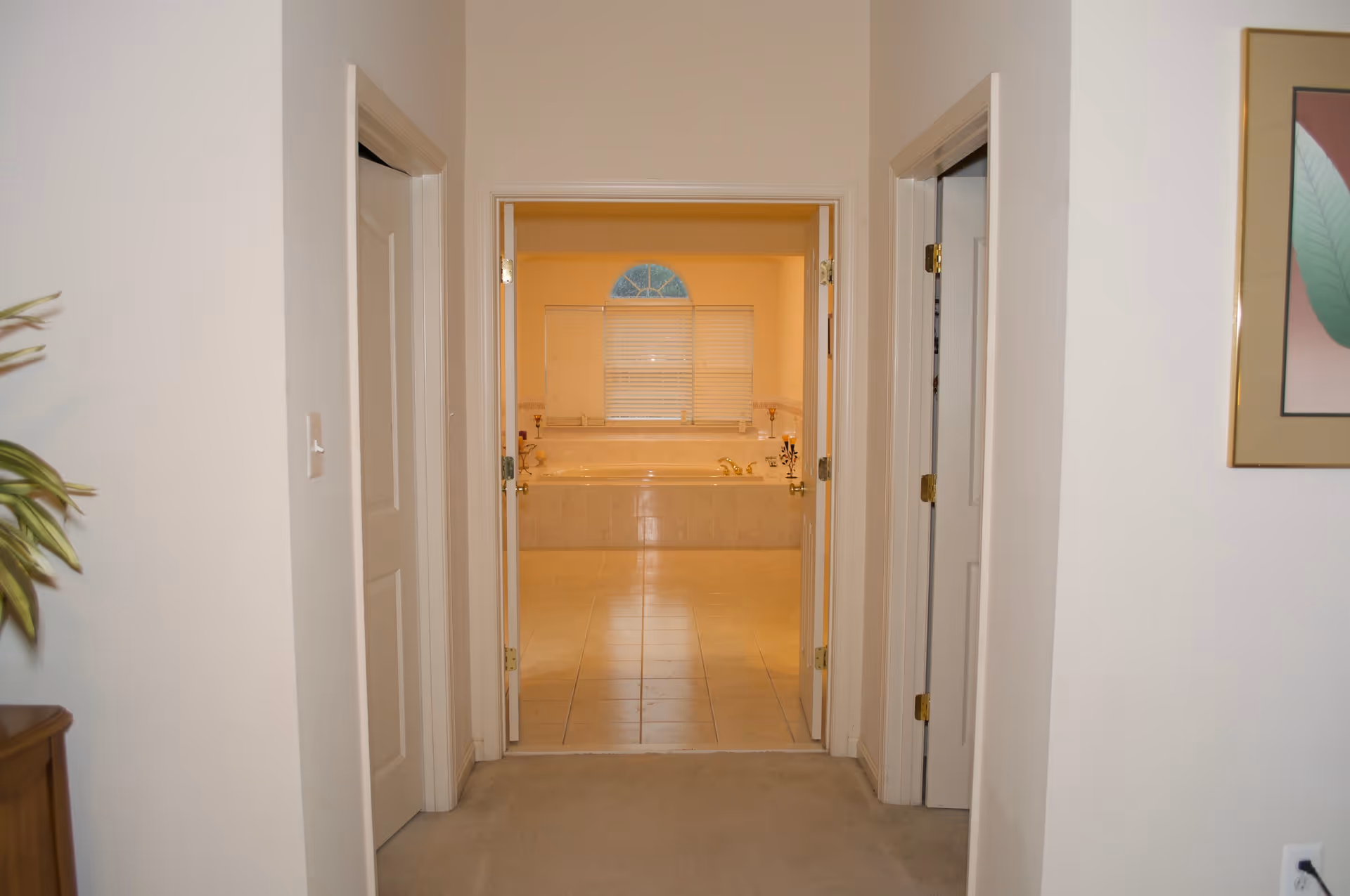 View down a hallway with beige carpet leading to a bathroom with tiled floor and a large bathtub under a window with blinds. There are two closed doors on either side of the hallway and a framed picture on the right wall.