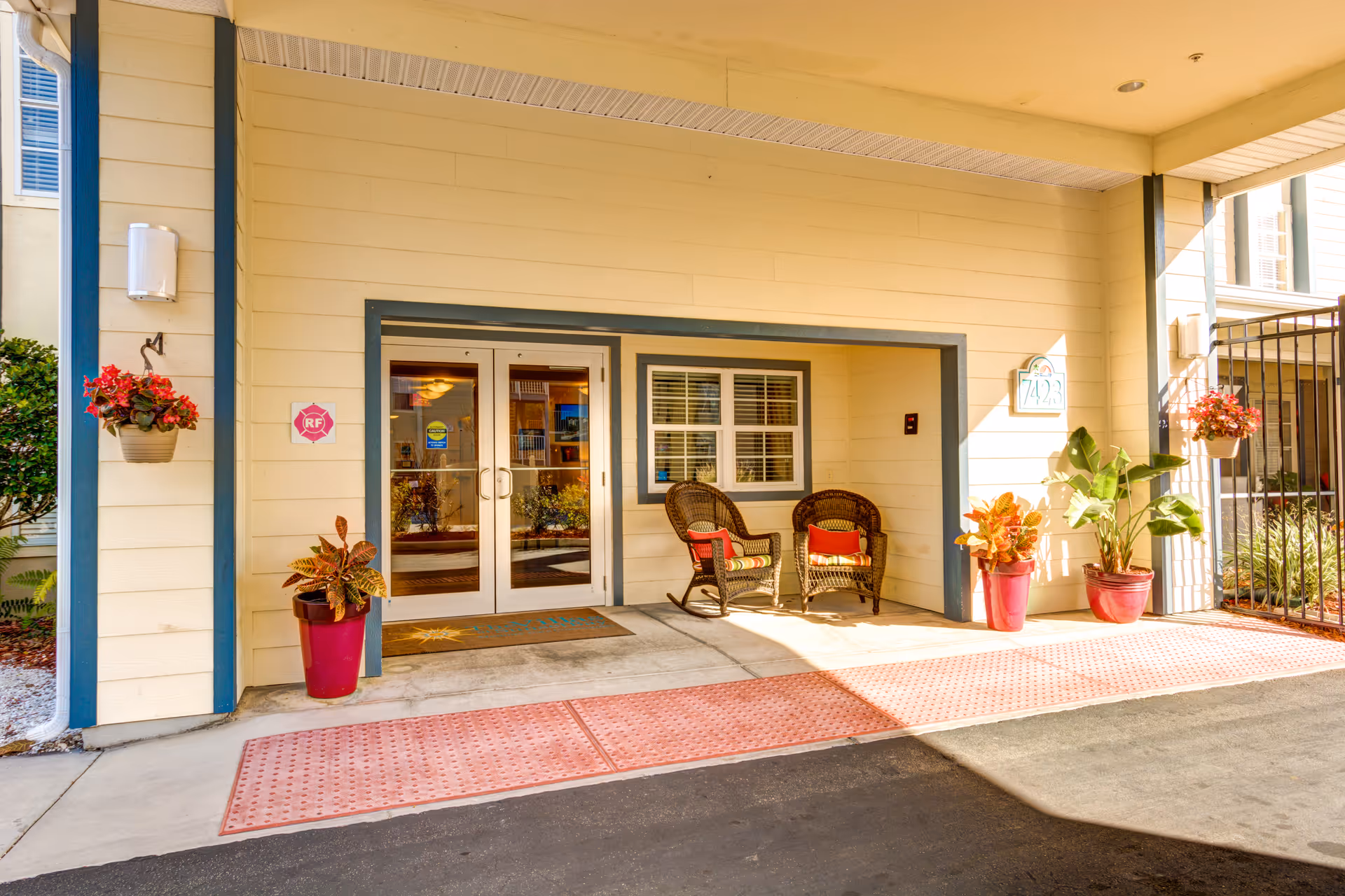 Covered entrance area of a senior living facility with double glass doors, two wicker chairs with cushions, potted plants, and hanging flower baskets. The building exterior is light yellow with blue trim, and there is a welcome mat in front of the doors.