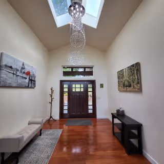 Interior entryway with a wooden front door featuring glass panels, a skylight above with a modern chandelier, a bench with a rug on the left side, a coat rack, and a black console table with decorative items on the right wall. Two paintings hang on the walls.