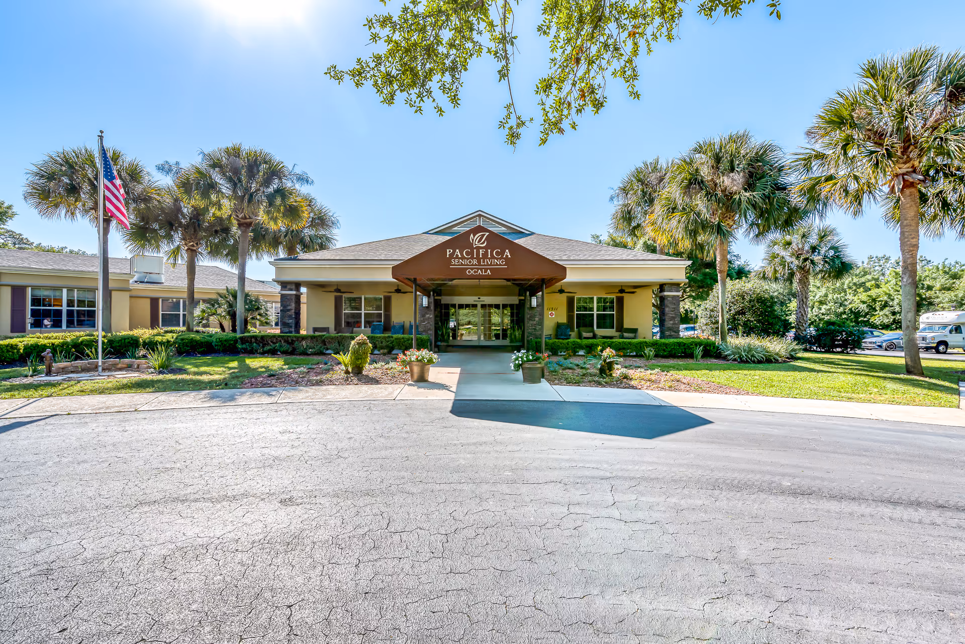 The front entrance of Pacifica Senior Living Ocala with a covered porte-cochere, palm trees, landscaping, and an American flag.