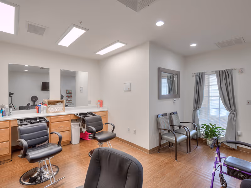 Interior of a salon area in a senior living facility with two black salon chairs in front of a counter with mirrors, various hair care products, and a basket of towels. There are additional chairs along the wall near a window with gray curtains and a green plant on the floor. The room has wood flooring and bright overhead lighting.