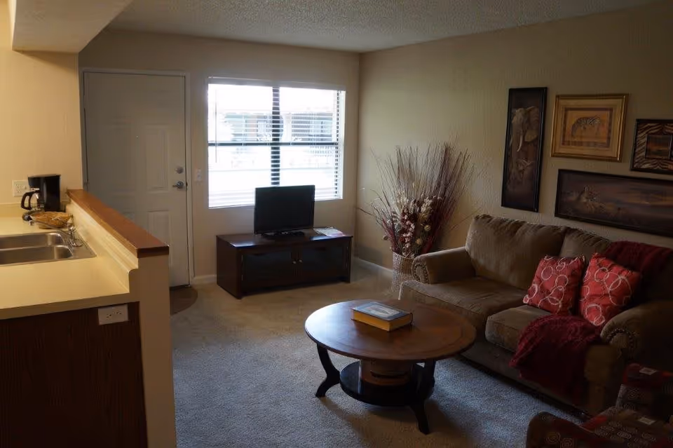 Living room with a sofa, round coffee table, small TV under a window, and a partial view of the kitchen counter.