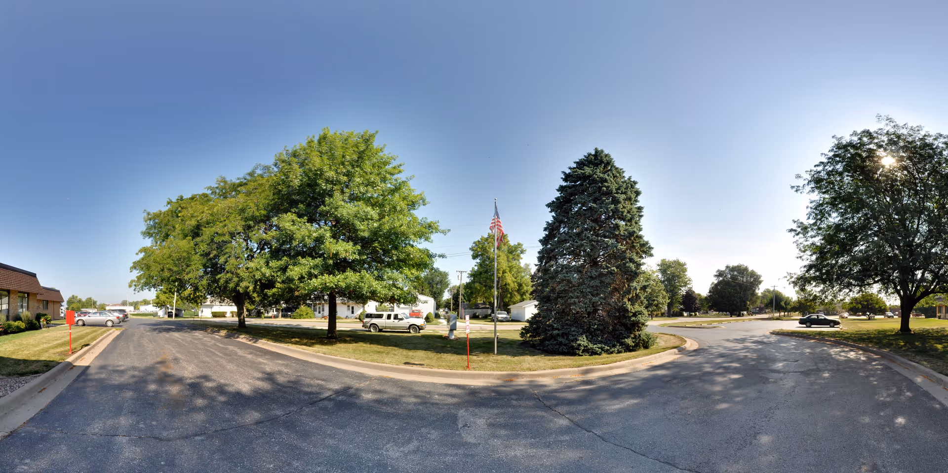 Driveway and landscaped front lawn with trees, a flagpole, and parked cars at the exterior of a senior living facility under a clear blue sky.