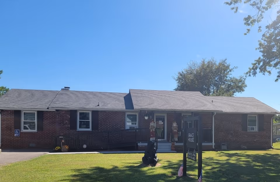 Single-story brick building with a ramp leading to the entrance, a sign in front reading 'Grace House Assisted Living for the Ages,' and a clear blue sky above.