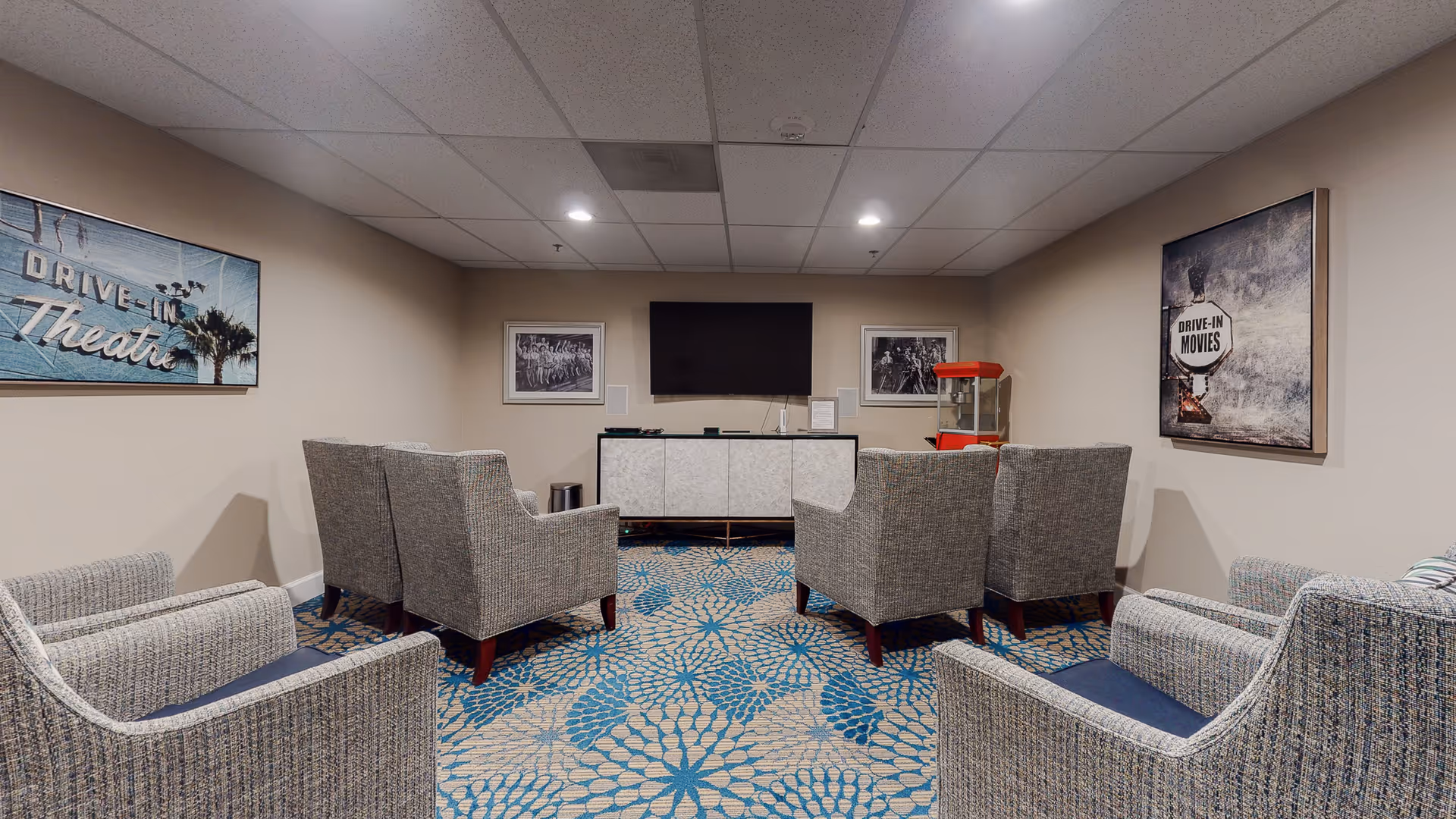 A small common/media room with upholstered armchairs arranged facing a wall-mounted TV, a popcorn machine, and decorative wall art.