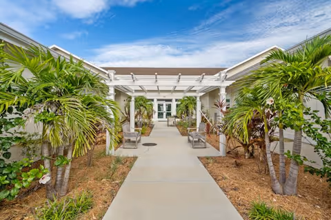 Outdoor walkway at Morningside House of Sarasota featuring a concrete path lined with palm trees and plants on both sides, leading to a white pergola and entrance doors under a partly cloudy blue sky.