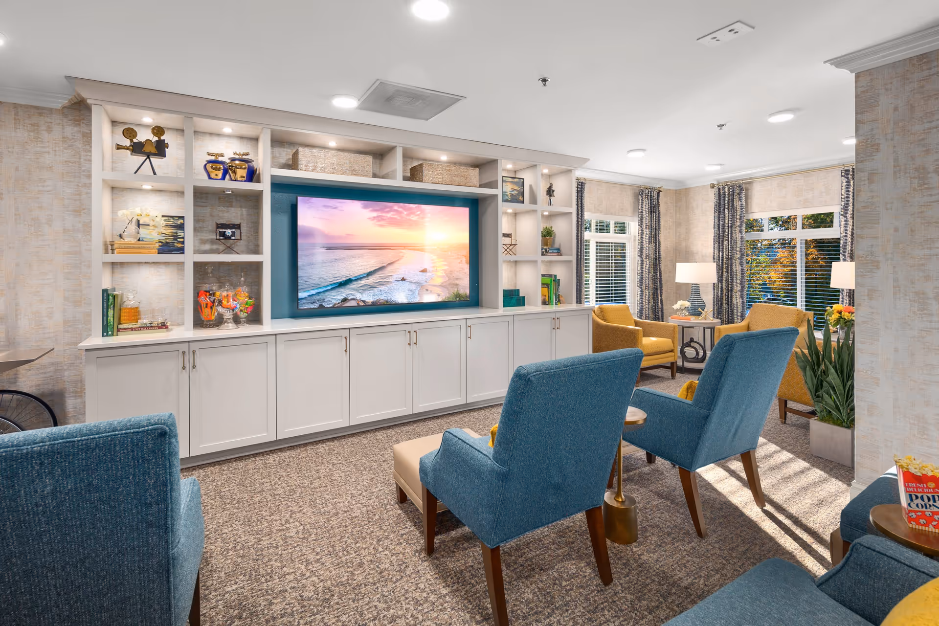 A cozy living room area with blue and yellow armchairs arranged around small tables. A large flat-screen TV is mounted on a white built-in shelving unit displaying a beach sunset scene. The room has large windows with patterned curtains, carpeted floor, and decorative items on the shelves including books, plants, and vintage cameras.