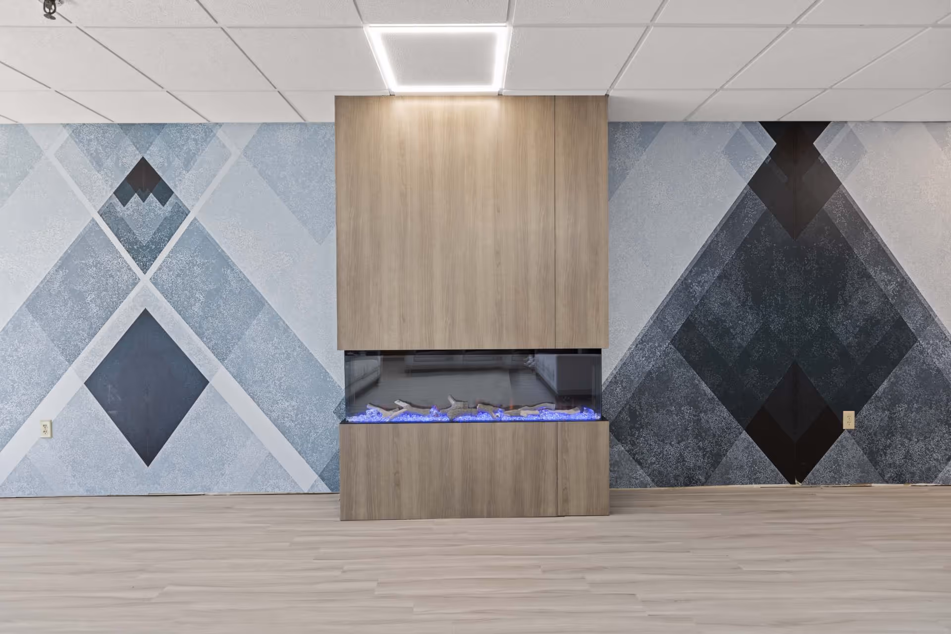 A modern electric fireplace with blue flames set in a wooden frame against a wall with geometric patterned wallpaper in shades of blue, gray, and black. The floor is light-colored wood, and the ceiling has white tiles with a square light fixture above the fireplace.