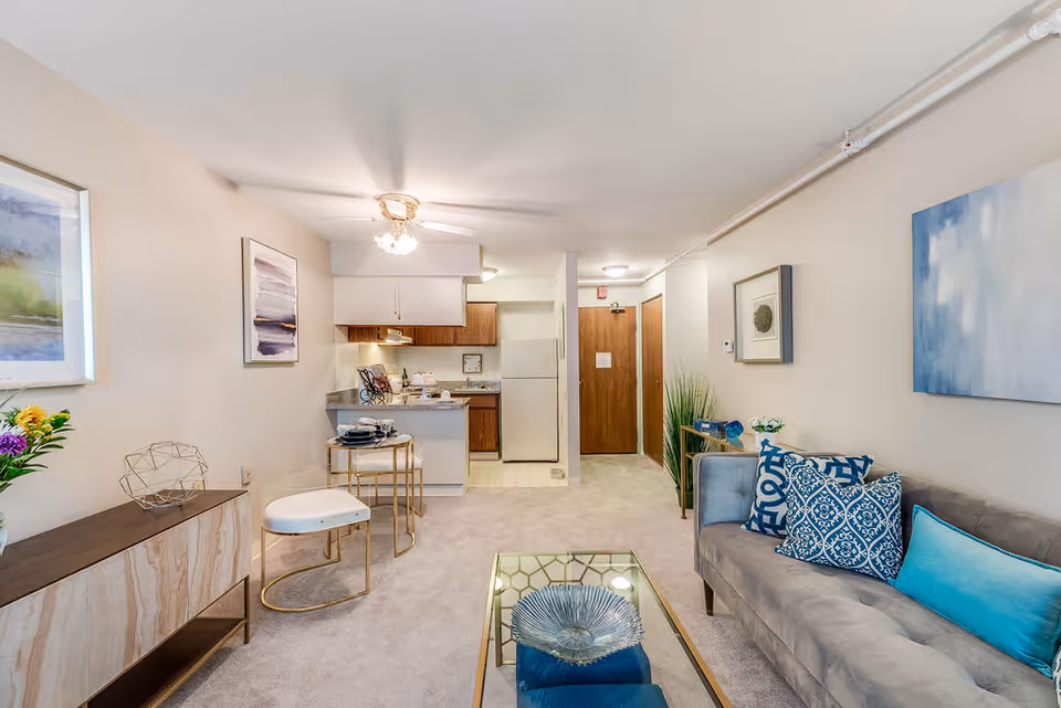 A bright and cozy living area in a senior living facility featuring a gray couch with blue and patterned pillows, a glass coffee table with a decorative bowl, a small dining table with two chairs, and a compact kitchen with wooden cabinets and white appliances. The walls are adorned with framed artwork, and there is a ceiling fan with lights. The entrance door is visible in the background.