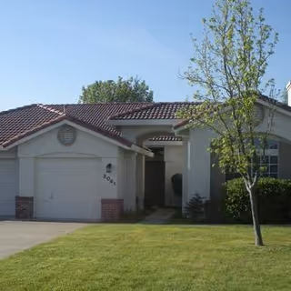 Single-story residential building with a tiled roof, attached garage, and a small tree in the front yard with green grass.