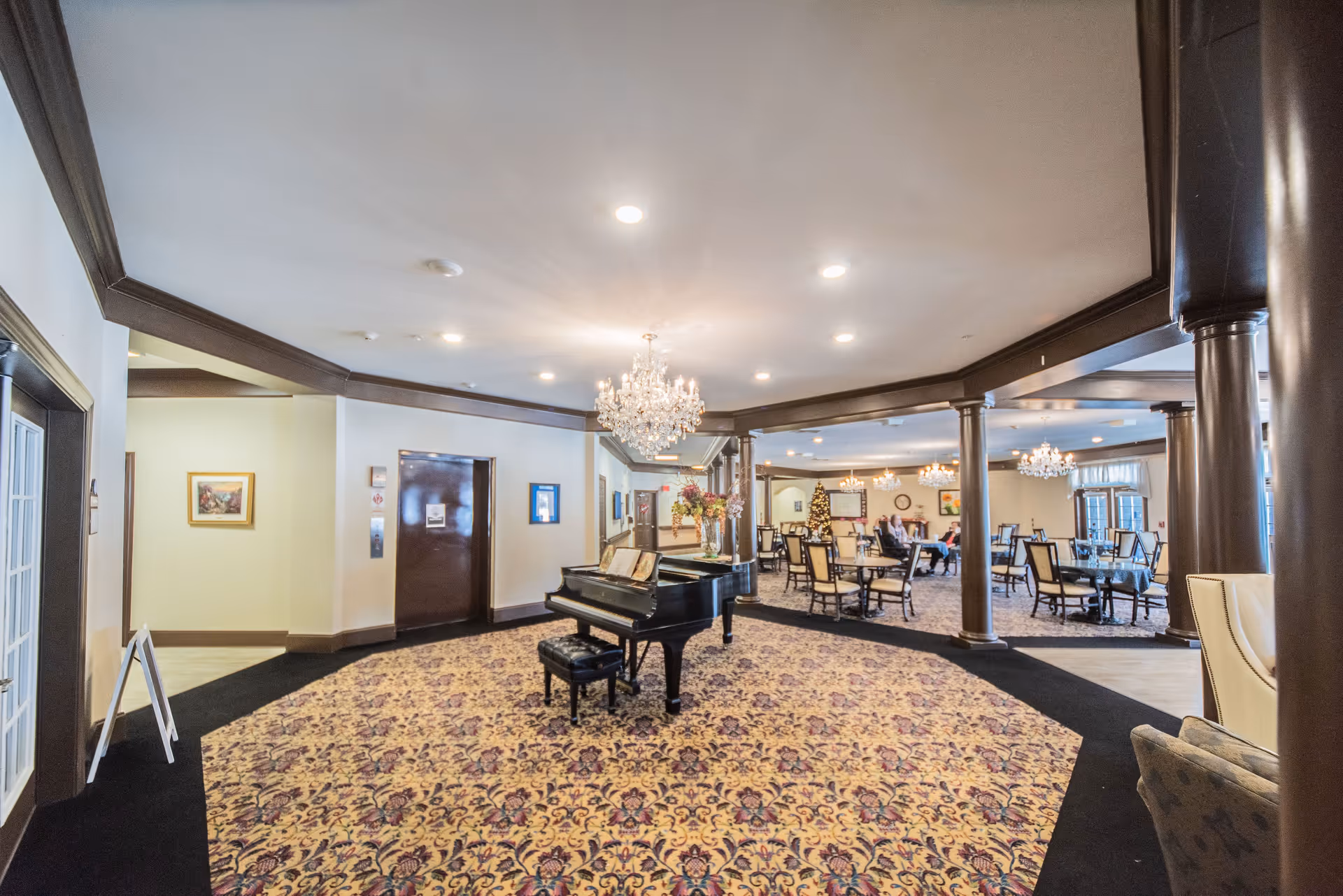 Interior view of a senior living facility common area featuring a grand piano on a patterned carpet, chandeliers hanging from the ceiling, and several tables and chairs arranged for dining or socializing. There are a few people seated in the background near a decorated Christmas tree.