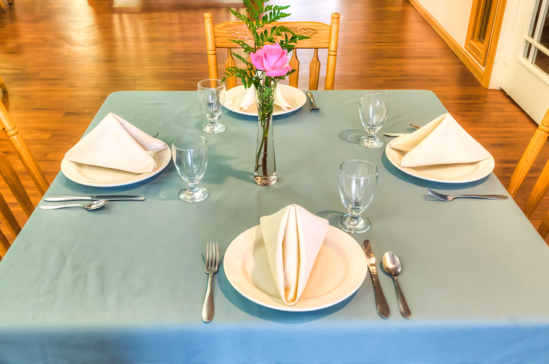 A dining table set for four with a light blue tablecloth, white plates, folded beige napkins, clear water glasses, and silverware. A small vase with a pink rose and green foliage is placed in the center of the table. The setting is in a room with wooden floors and wooden chairs.