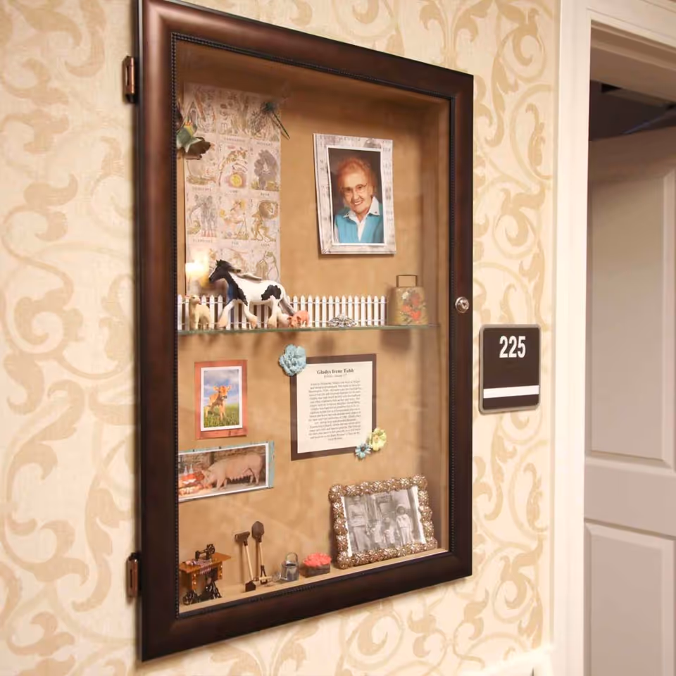 A display case mounted on a patterned beige wall next to a door labeled 225. Inside the case are various small items including a framed photo of an elderly woman, miniature animal figurines, a small white picket fence, a framed black and white photo, and other decorative objects.