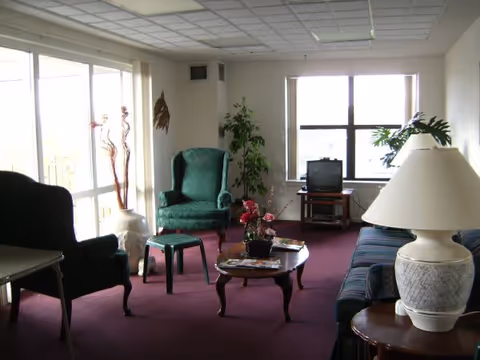 Small communal living room with upholstered chairs and a sofa around a coffee table, a TV on a stand, large windows, and potted plants.