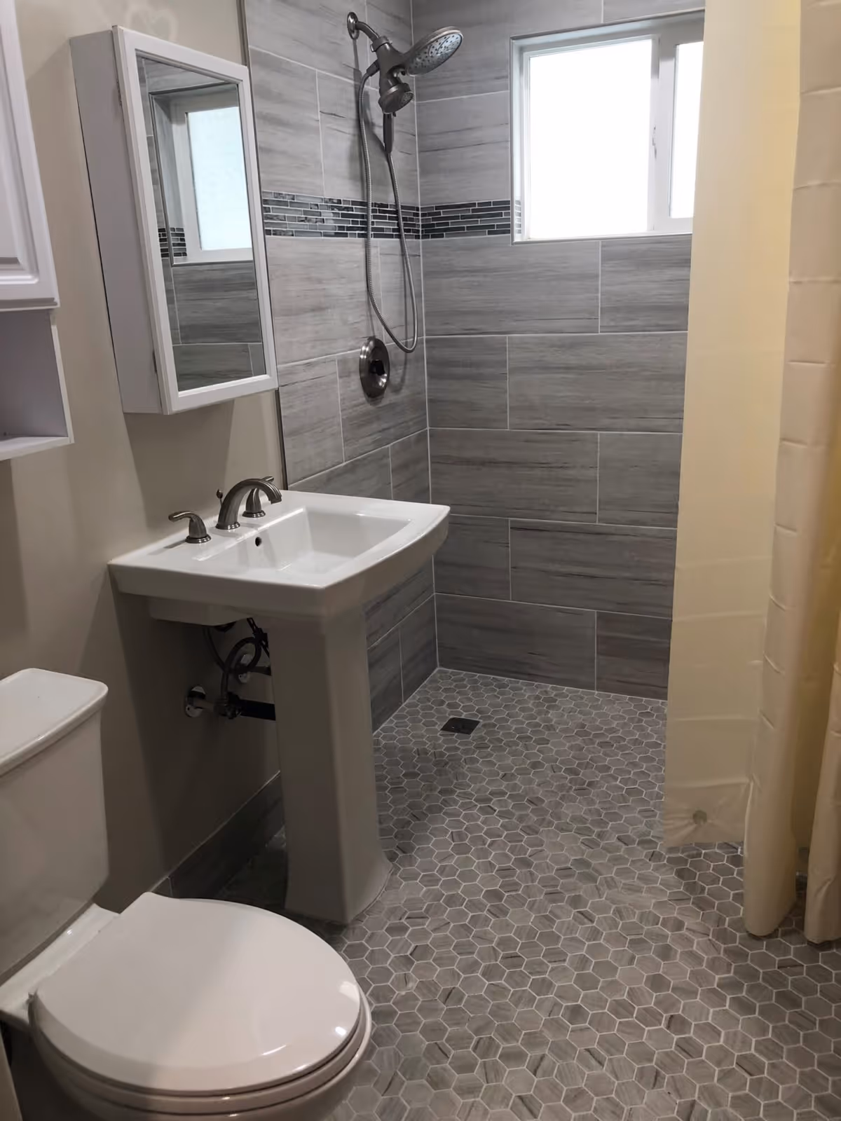 A modern bathroom with a white toilet, a pedestal sink with chrome faucets, a wall-mounted mirrored medicine cabinet, and a walk-in shower with gray tiles and a rainfall showerhead. The floor has hexagonal gray tiles and there is a small window in the shower area.