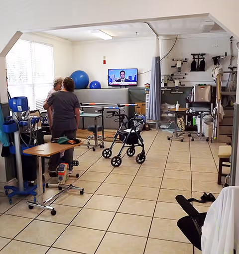 A rehabilitation/therapy room with walkers, exercise balls, parallel bars, and two people near a wall-mounted TV.