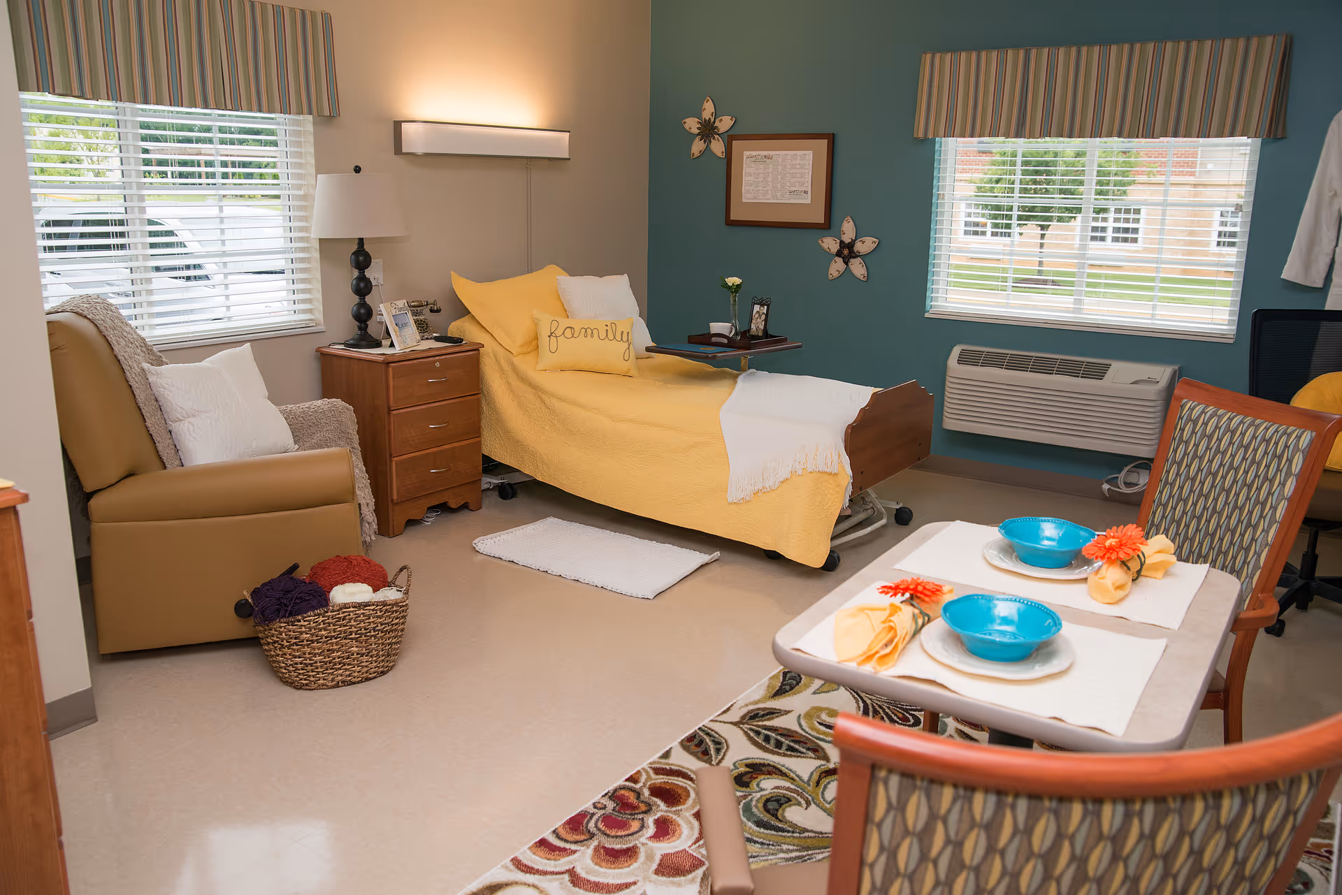 A cozy senior living room with a single bed covered in a yellow blanket and a pillow with the word 'family'. There is a brown armchair with a white pillow and a basket of yarn next to it. A small wooden nightstand with a lamp and picture frame is beside the bed. Two windows with striped valances let in natural light. A small dining table set for two with blue bowls, yellow napkins, and orange flowers is in the foreground. The walls are painted teal and beige, and there is a wall-mounted air conditioner beneath one window.