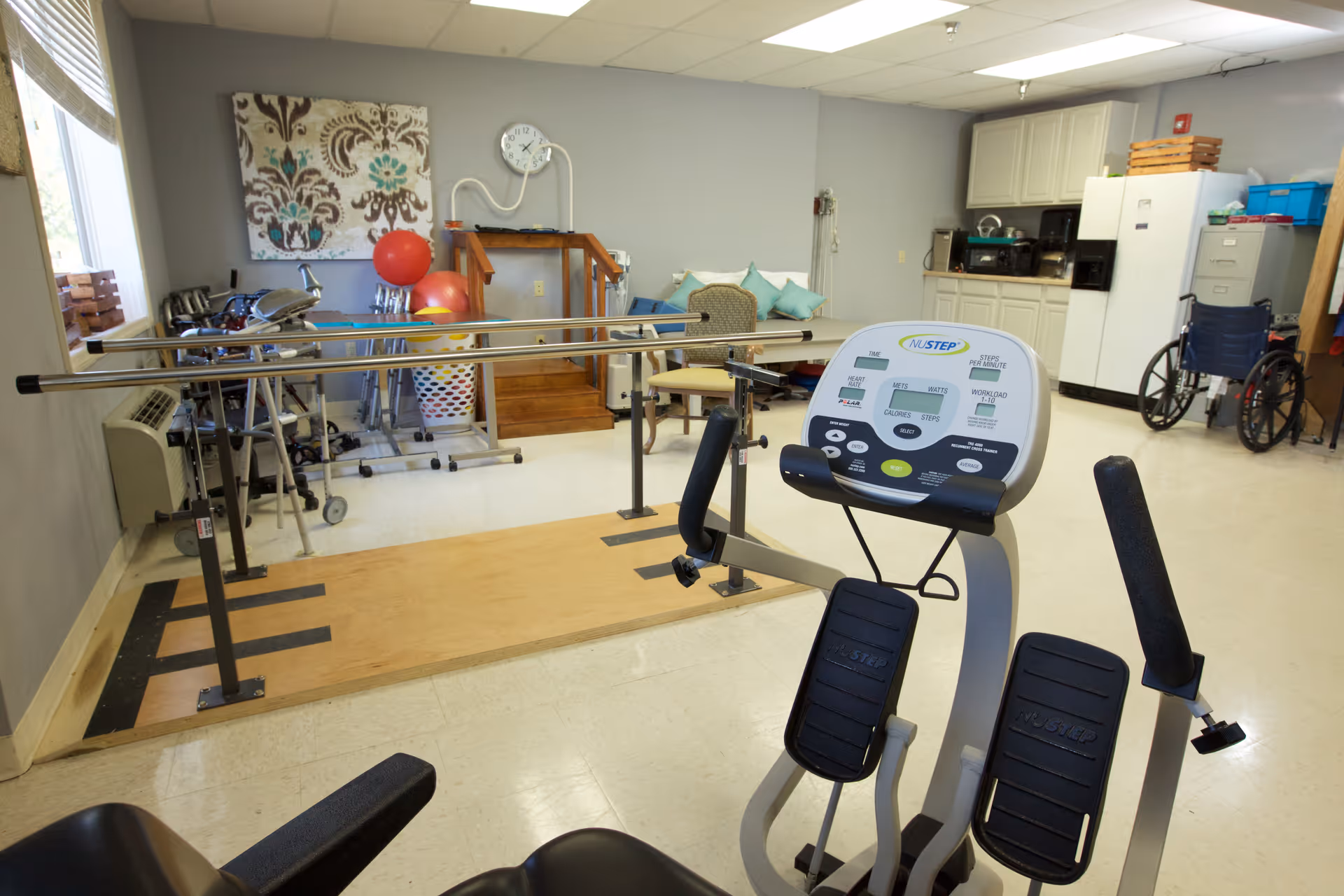 A rehabilitation or physical therapy room in a senior living facility with exercise equipment including a NuStep machine, parallel bars for walking practice, therapy balls, a wheelchair, and a small seating area with chairs and cushions. The room also has a kitchenette area with cabinets, a refrigerator, and a filing cabinet.