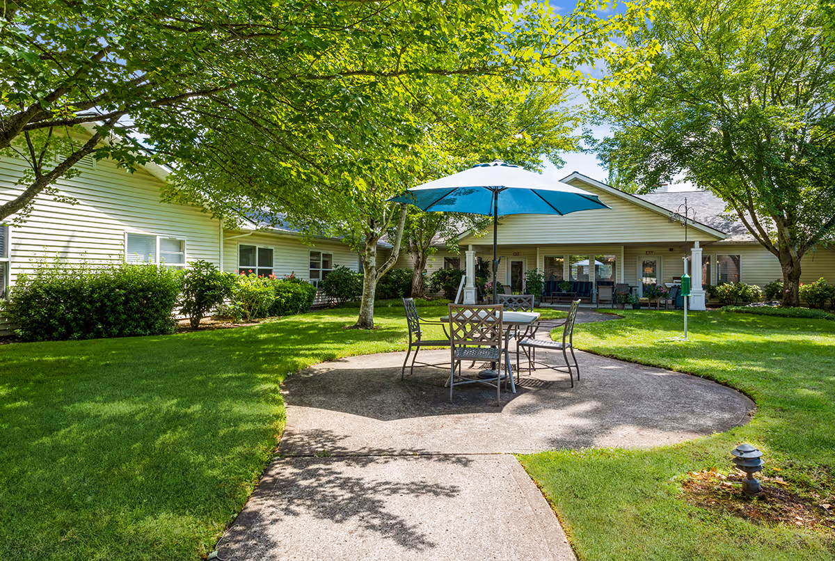 Shaded courtyard with a round table, chairs and a blue umbrella on a concrete patio in front of a single-story residential building surrounded by trees and lawn.