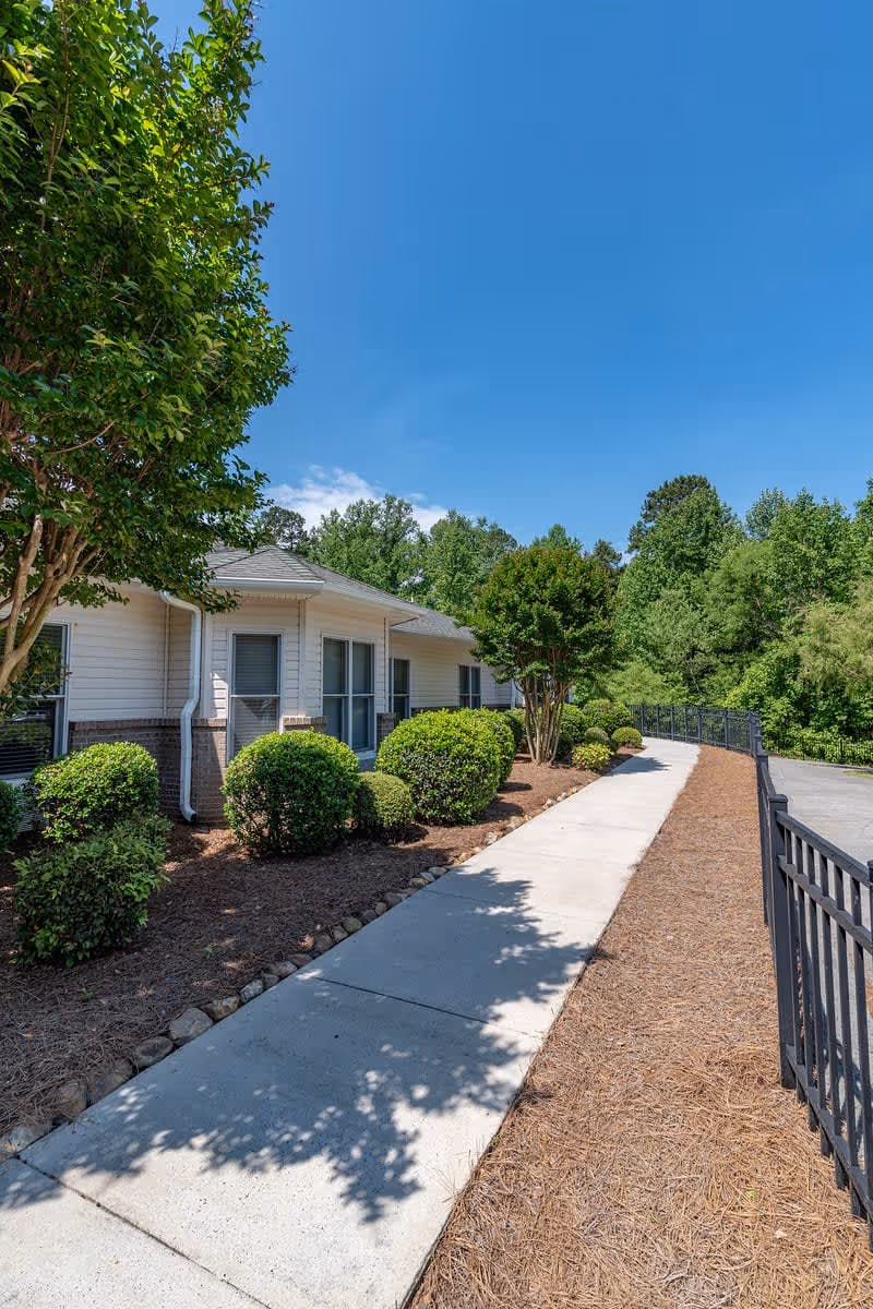 Concrete sidewalk lined with shrubs and a fence leading past a single-story building under a clear blue sky.