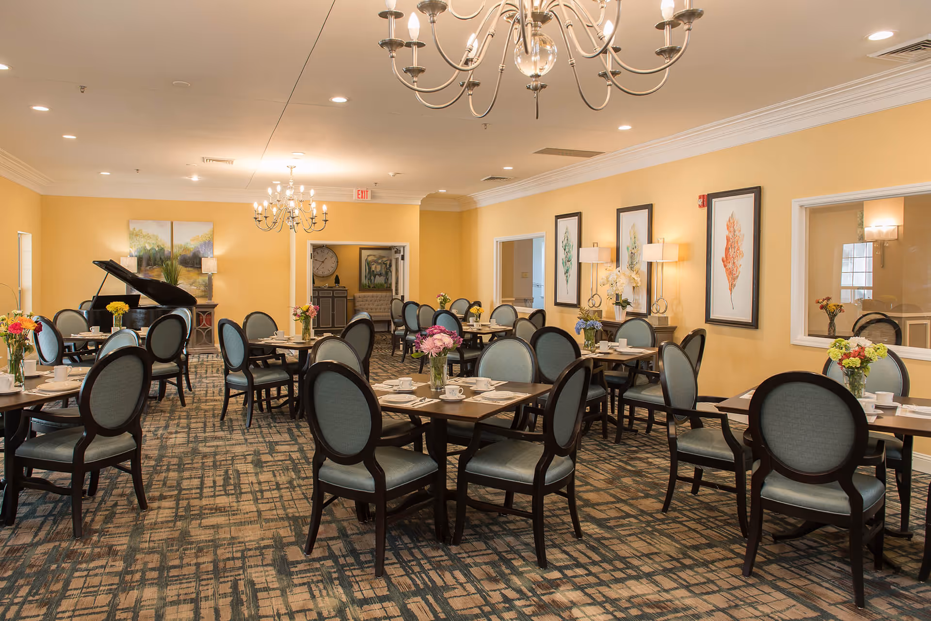 A spacious dining room with multiple tables and chairs arranged neatly. Each table has a vase with colorful flowers and place settings with cups and saucers. The room features yellow walls, framed botanical artwork, chandeliers, and a grand piano in the corner.