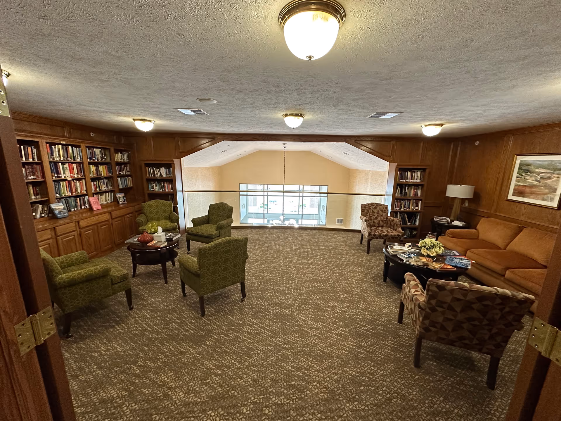 Comfortable lounge/library with bookshelves, green armchairs, a sofa, and coffee tables overlooking an interior balcony.