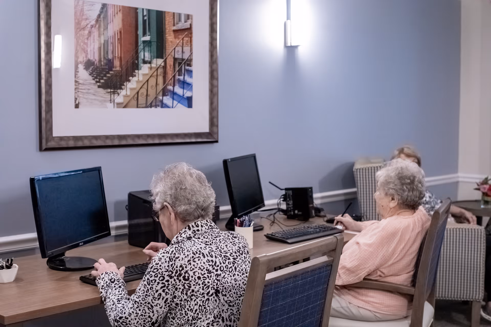Two elderly women sitting at a desk using desktop computers in a room with light blue walls, a framed picture of colorful stairs on the wall, and a third person sitting in a chair in the background.
