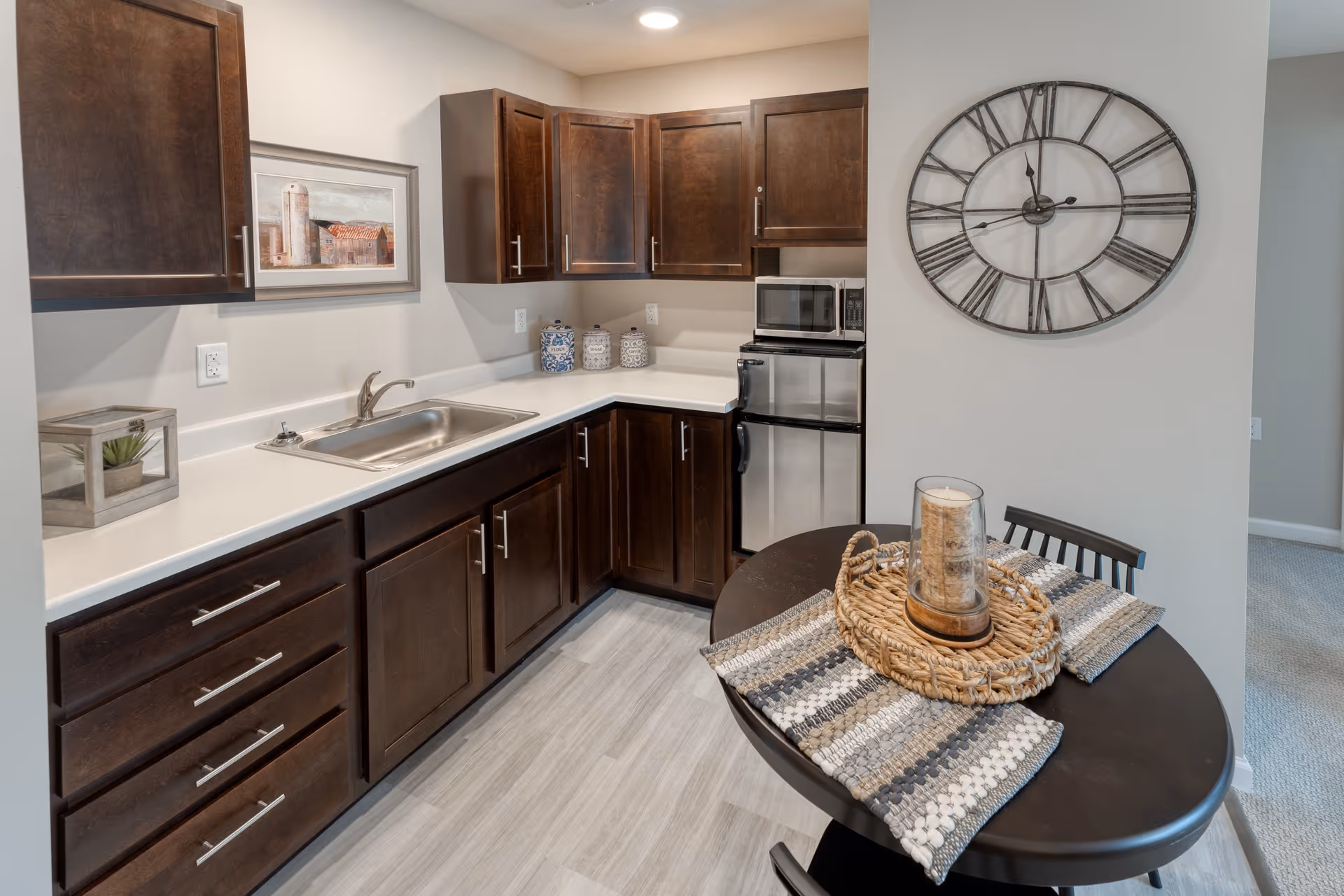A compact kitchen area with dark wood cabinets, a stainless steel sink, a small refrigerator, and a microwave. A round dining table with two chairs is set with woven placemats and a decorative candle centerpiece. A large wall clock with Roman numerals hangs on the adjacent wall, and a framed picture of a barn is mounted above the countertop.