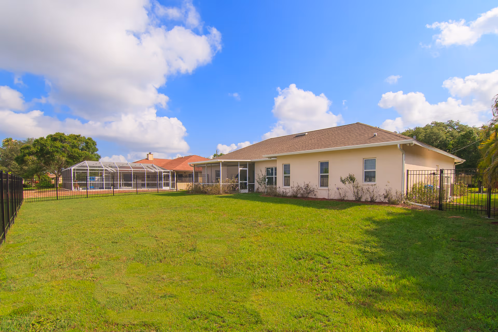 A single-story beige building with a brown roof surrounded by a green grassy yard under a partly cloudy blue sky. There is a black metal fence enclosing the yard, and another house with a screened pool enclosure is visible in the background.