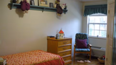 A small bedroom with a single bed covered in a red and orange patterned bedspread. There is a wooden dresser with a small decorative house and other items on top. A cushioned chair with a purple pillow is placed near a window with blue valance curtains. On the wall above the bed is a shelf holding framed photos and stuffed animals. The floor is wooden, and slippers are placed near the chair.