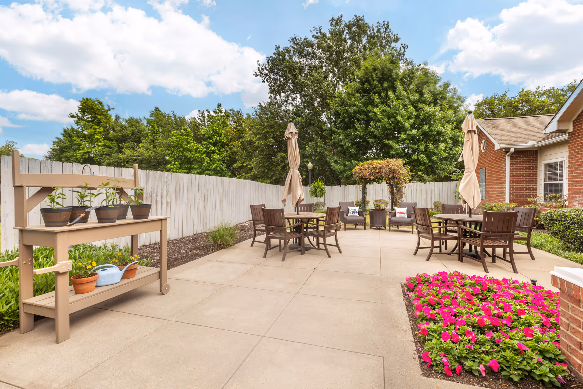 Outdoor patio area with several brown tables and chairs, each table having a closed beige umbrella. There is a wooden potting bench with potted plants and gardening tools on the left side. The patio is surrounded by a white wooden fence, green trees, and a flower bed with bright pink flowers on the right side near a brick building.