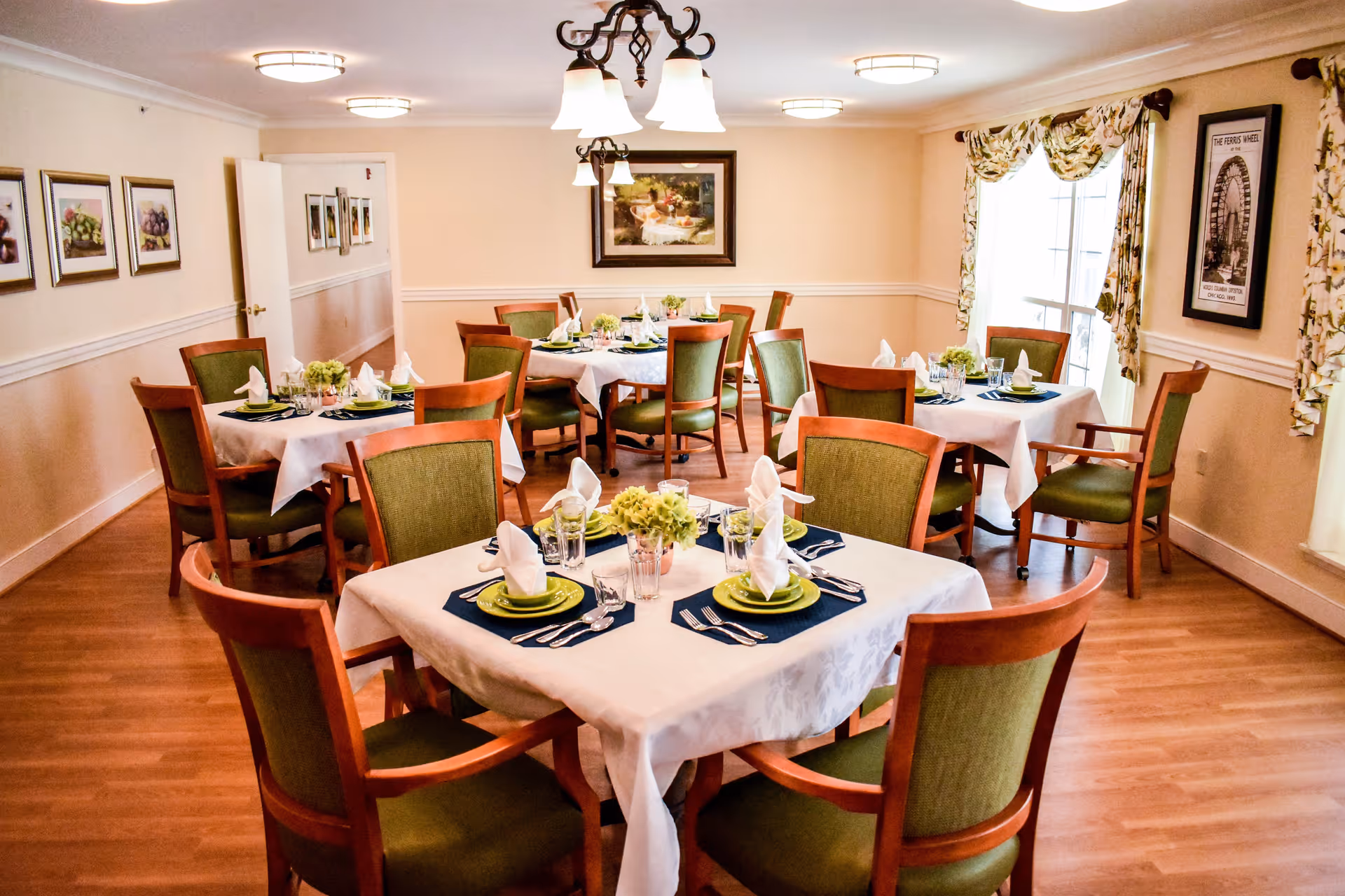 A bright dining room with several square tables covered with white tablecloths, each set with green plates, silverware, glasses, and napkins folded upright. The chairs have wooden frames and green upholstery. The room has light-colored walls adorned with framed pictures and windows with floral curtains, and wooden flooring.