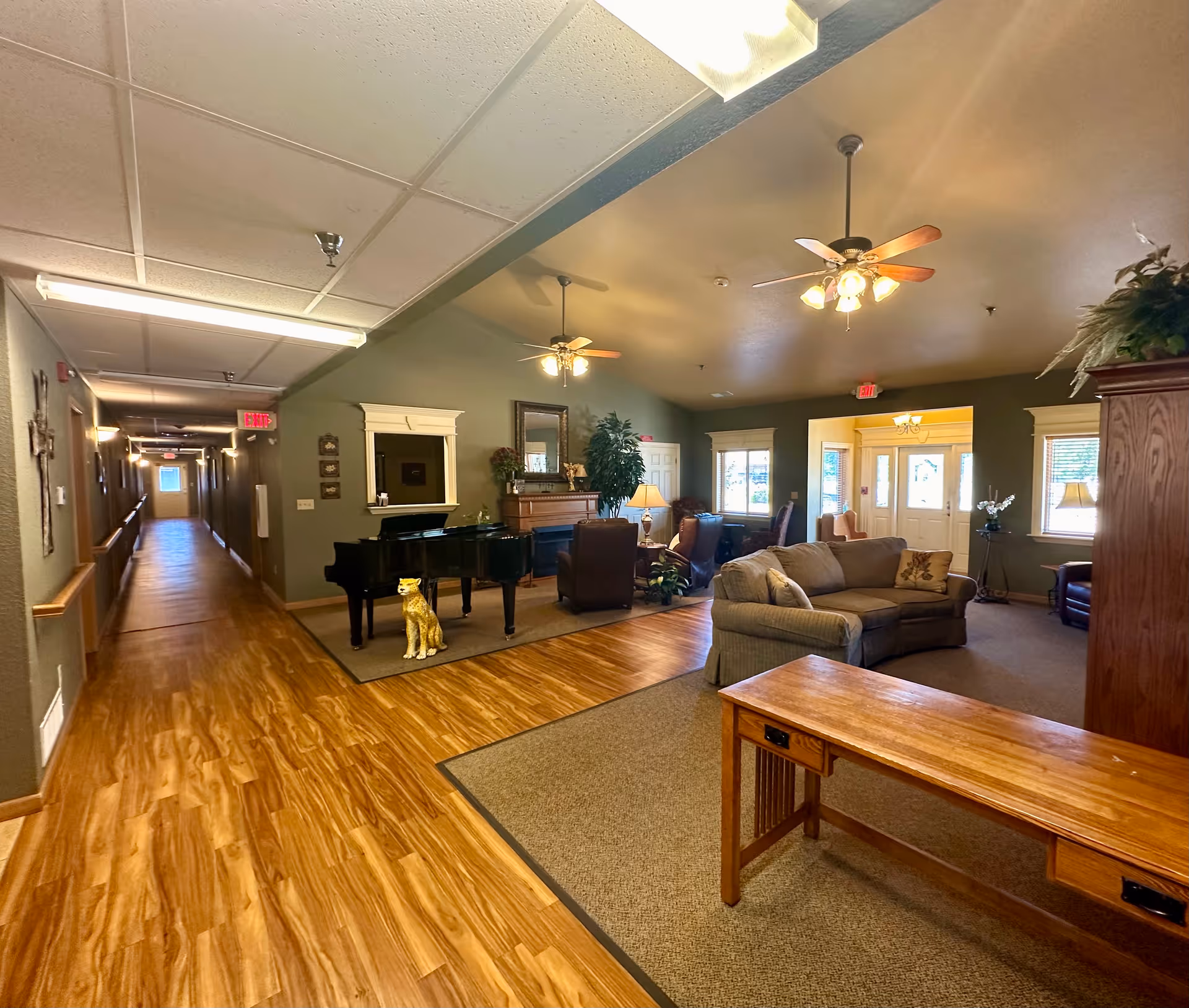 Interior view of a senior living facility lounge area with wooden flooring and a long hallway on the left. The lounge features a grand piano with a decorative cat statue in front, comfortable armchairs, a sofa, ceiling fans with lights, and large windows letting in natural light. There is a wooden table in the foreground and plants and lamps adding to the cozy atmosphere.