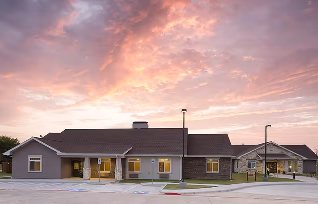 Exterior view of a single-story senior living facility building with a combination of stone and siding facade under a colorful sunset sky with pink and orange clouds. The building has multiple windows and two street lamps in front, with a parking area including handicap parking spaces.