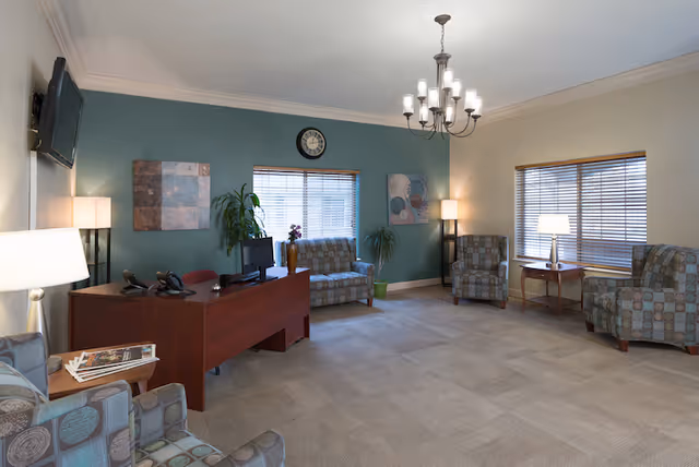 A well-lit living room area with beige carpet and walls painted in beige and teal. The room features a wooden desk with two telephones and a computer monitor, several patterned armchairs, a loveseat, two windows with blinds, two floor lamps, a table lamp, wall art, a clock, and some potted plants. A chandelier hangs from the ceiling.