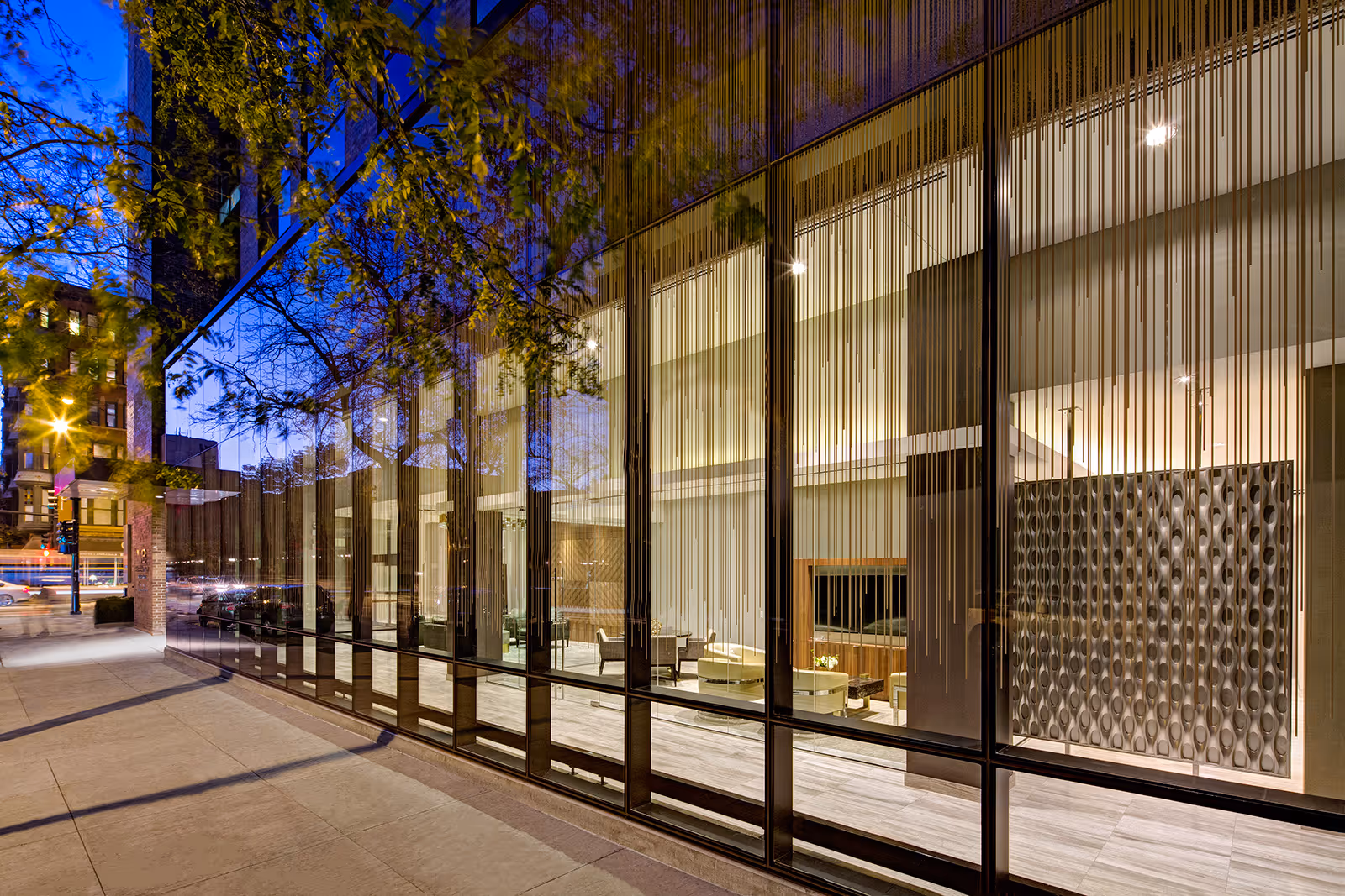 Glass-front modern building at dusk showing a well-lit interior lounge visible through tall windows.