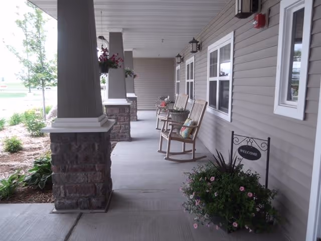 Covered porch area with several wooden rocking chairs, potted plants, and hanging flower baskets along the side of a building with beige siding and white-trimmed windows. A small decorative sign reading 'WELCOME' is placed among the plants near the entrance.