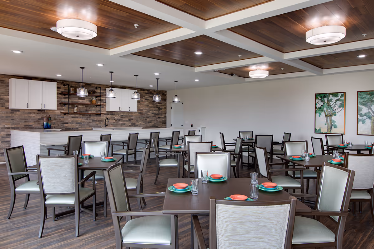 A modern dining room in a senior living facility with multiple tables and chairs arranged neatly. Each table is set with plates, bowls, and glasses. The room features a wooden floor, a ceiling with wooden panels and recessed lighting, and a brick accent wall with white cabinets and hanging pendant lights above a counter. Two framed paintings of trees hang on a white wall.