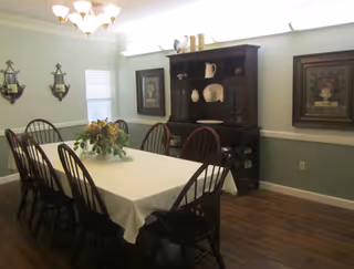 A dining room with a long rectangular table covered with a white tablecloth and surrounded by eight wooden chairs. A floral centerpiece is on the table. The room has light green walls with white trim, two framed paintings, a dark wooden hutch with decorative items, and two wall-mounted candle holders. A chandelier with multiple lights hangs above the table.
