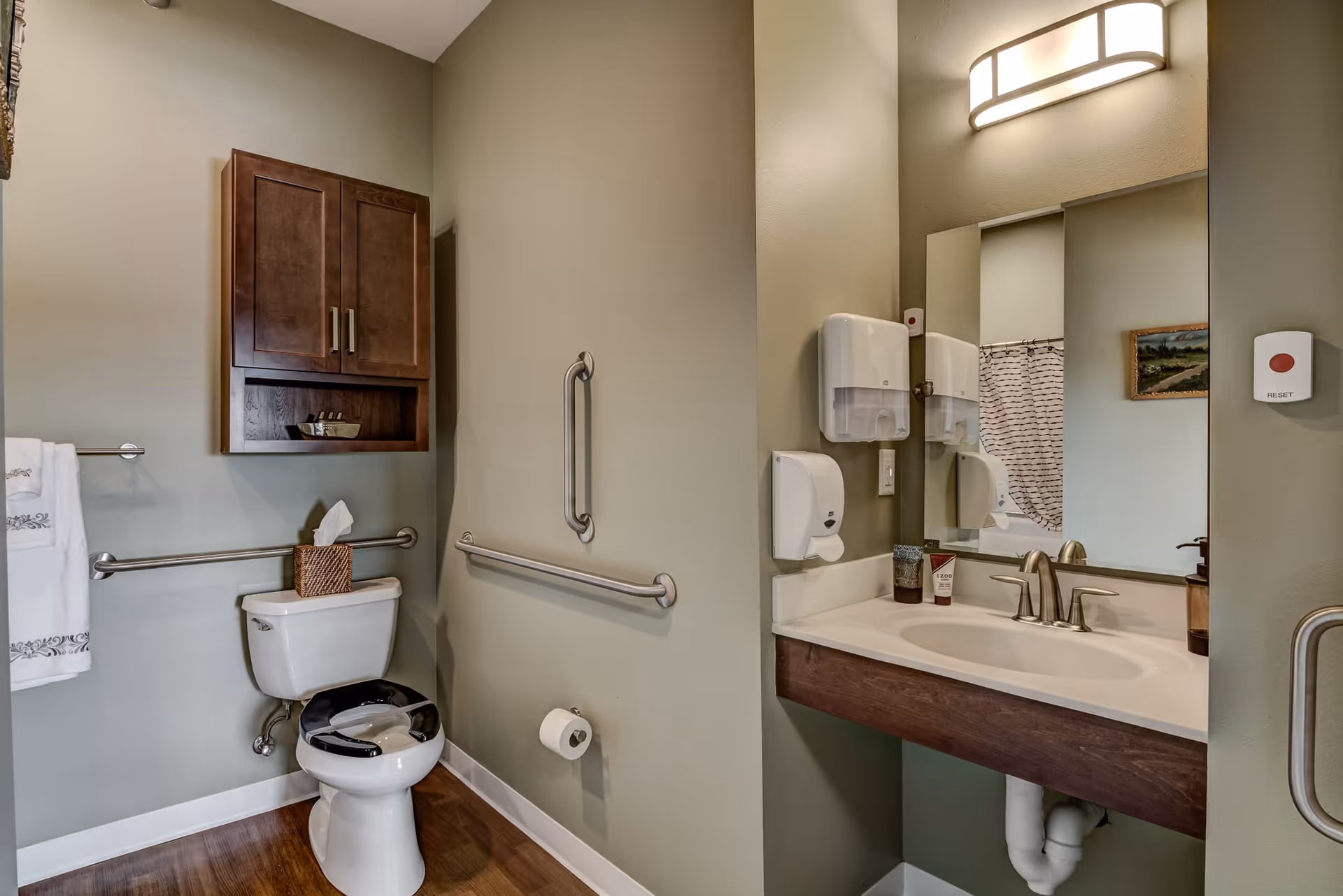A clean and accessible bathroom with a toilet featuring a raised seat and safety grab bars on the walls. Above the toilet is a wooden cabinet. To the right is a sink with a large mirror, soap dispenser, and a wall-mounted paper towel dispenser. The walls are painted light green and the floor has wood-like flooring.