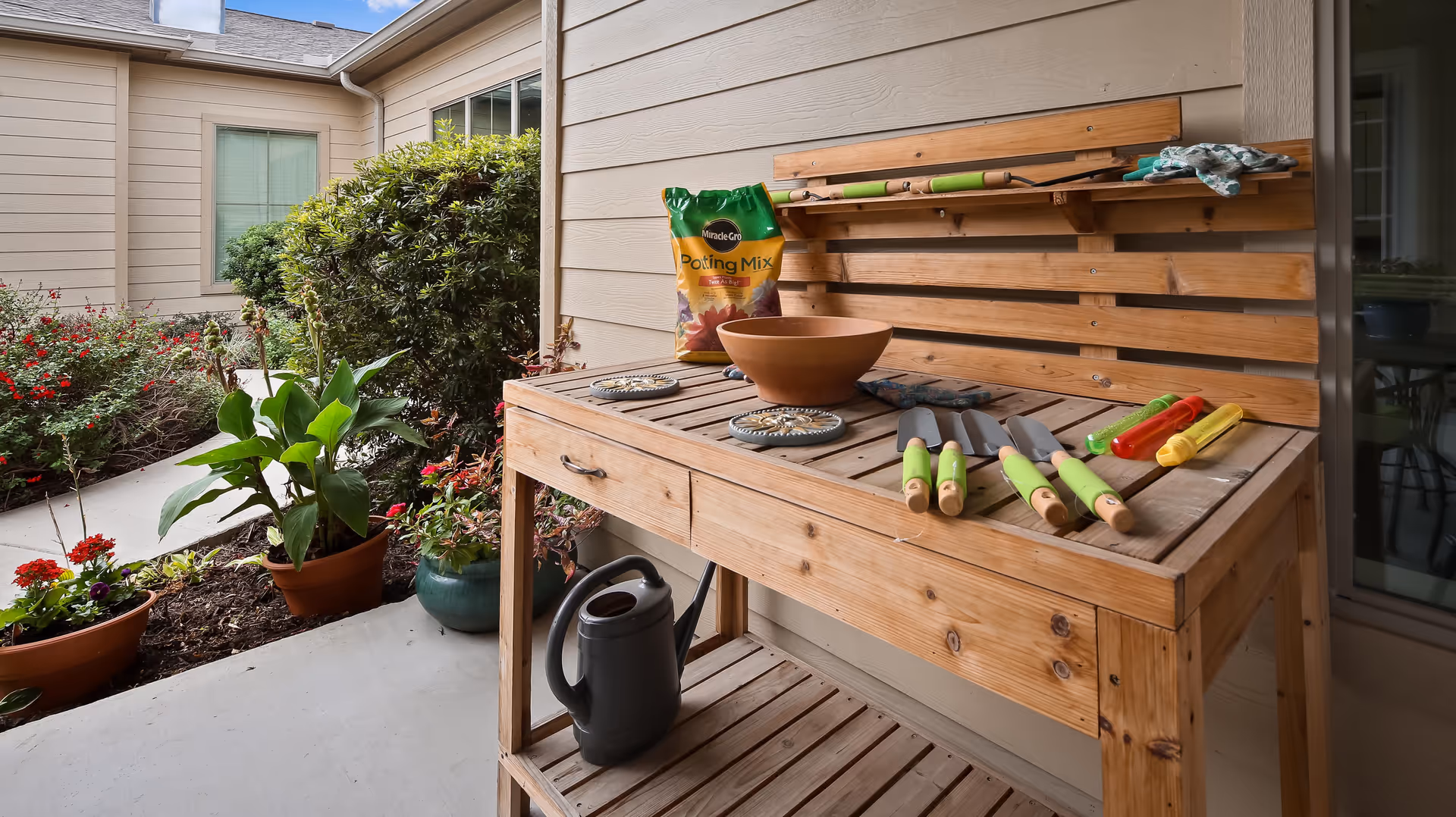 Outdoor gardening workspace with a wooden potting bench holding gardening tools, a bag of potting mix, a watering can, and a clay pot. Surrounding the area are potted plants and bushes near the exterior wall of a building.