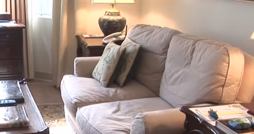 A cozy living room area with a beige sofa adorned with two decorative pillows. Next to the sofa is a wooden side table with a decorative lamp and a telephone. A window with light curtains allows natural light to brighten the room. In front of the sofa, there is a wooden coffee table with some items on it.